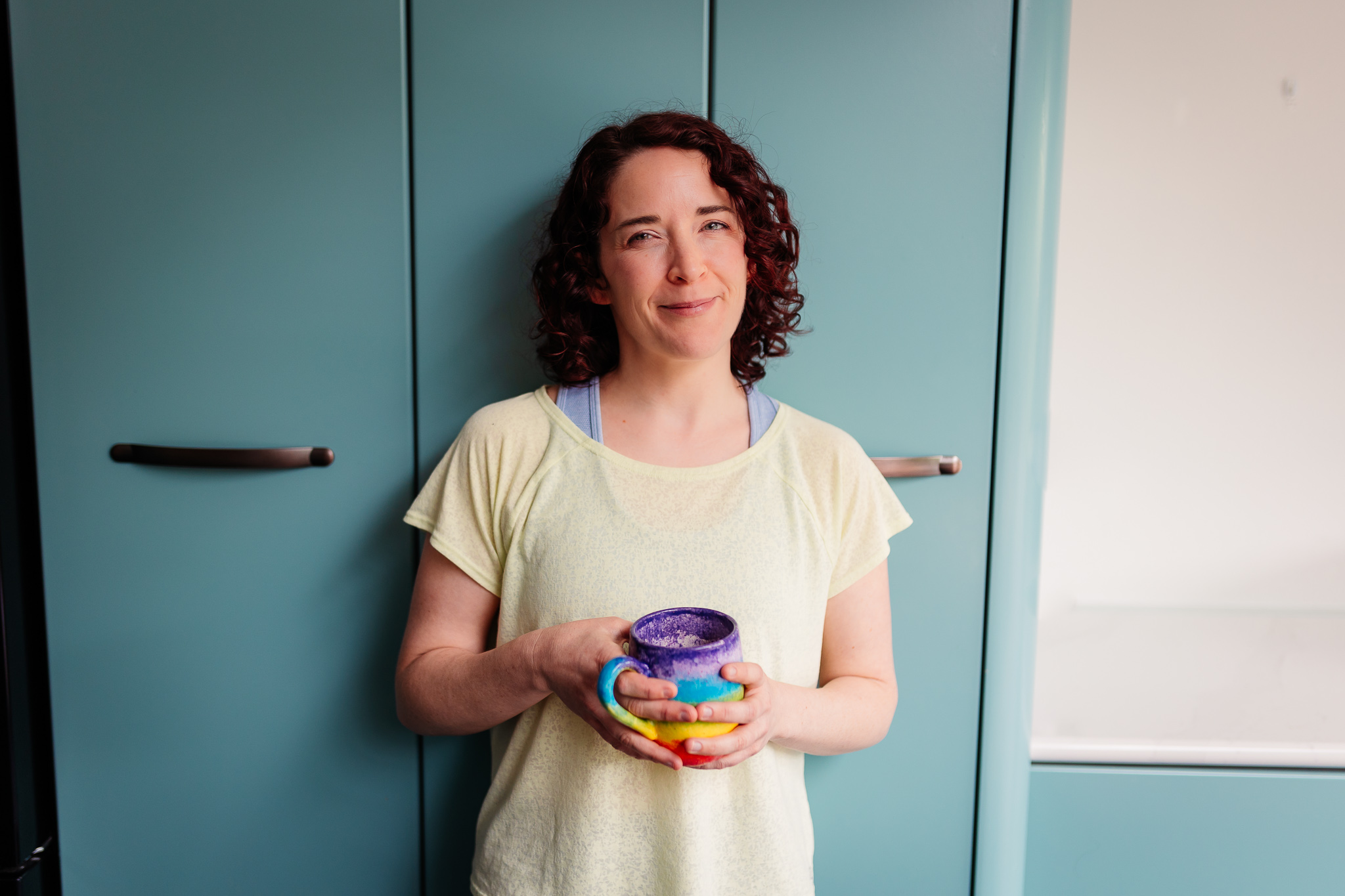 Emmeline Kemp the founder of Green Room Health: A Pilates and Barre Studio in Danbury, Essex. Emmeline is smiling, leaning against a green cupboard, whilst holding a rainbow coffee cup.