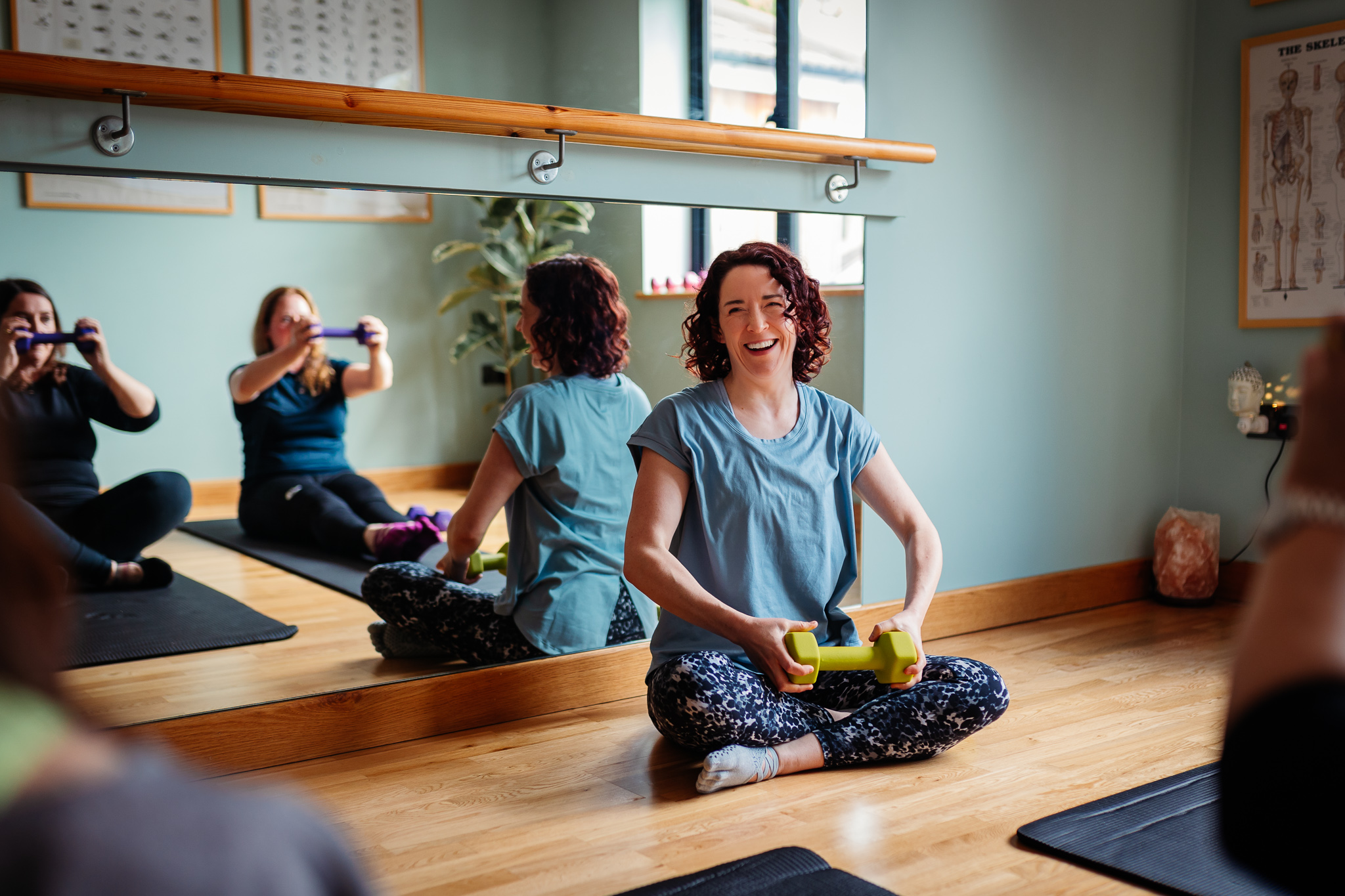 Three women are smiling and sitting on Pilates mats in front of studio owner Emmeline Kemp whilst holding hand weights in the studio at Green Room Health in Danbury, Essex.